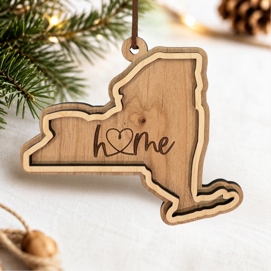 Layered New York wood ornament featuring engraved “home” script and faux leather hanger on a soft neutral holiday background with greenery and warm lights.