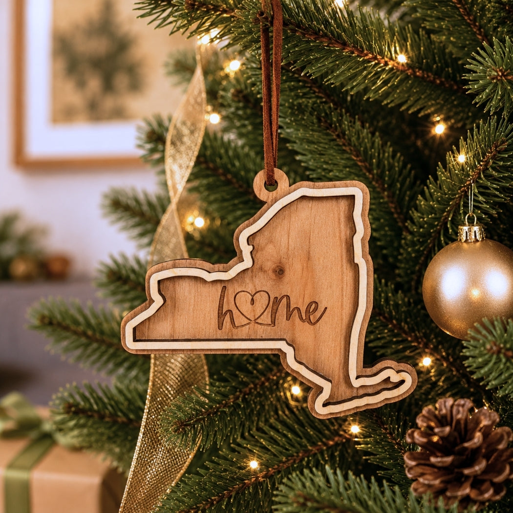 Layered New York “home” ornament hanging on a decorated Christmas tree with warm white lights, pinecones, and gold ornaments.
