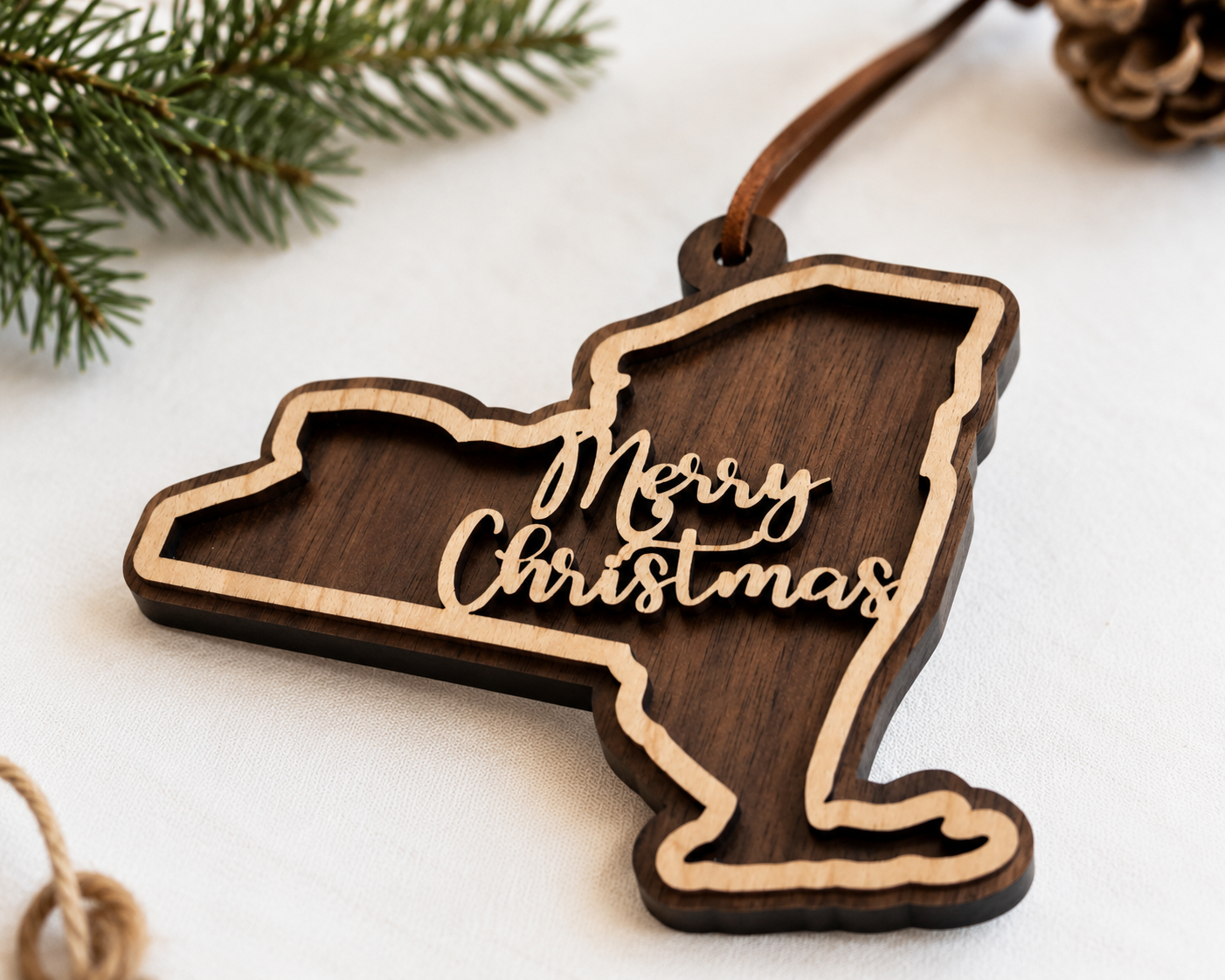 Layered New York state Christmas ornament in walnut and maple plywood with “Merry Christmas” script, photographed at an angled close-up with evergreen branches and holiday styling.