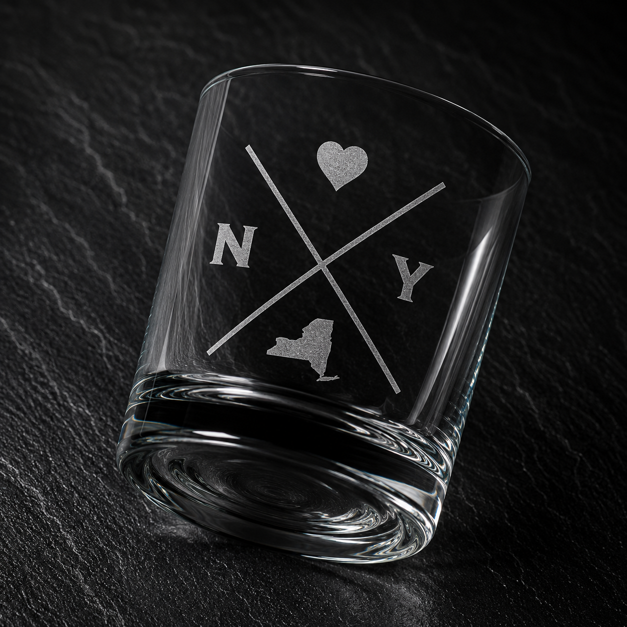 Angled engraved New York whiskey glass featuring frosted New York silhouette and modern crossed-letter design on a dark textured surface.