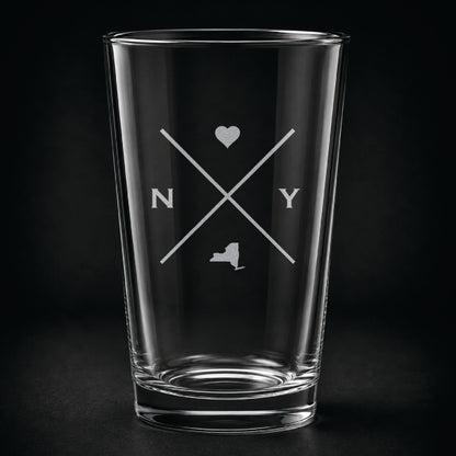 Engraved New York pint glass featuring a minimalist crossed-line design with heart detail and New York silhouette on a dark background.