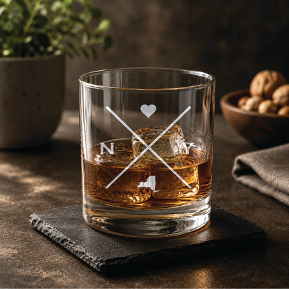 Engraved New York rocks glass with crossed-line monogram design, heart detail, and New York state silhouette, styled with whiskey and ice on a slate coaster in a warm, upscale bar setting.