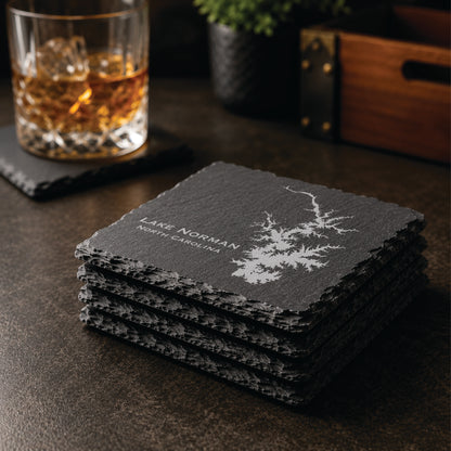 Stack of engraved Lake Norman slate coasters in a warm bar-style setting with whiskey glass nearby, featuring a detailed lake map design and North Carolina text.