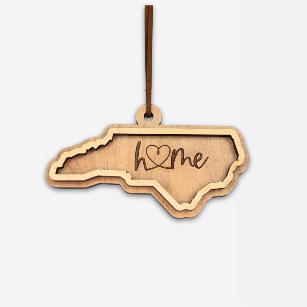 Layered North Carolina wood ornament with engraved “home” script and faux leather cord on white background.