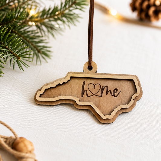 Layered North Carolina wood ornament featuring engraved “home” script and faux leather hanger on a soft neutral holiday background with greenery and warm lights.