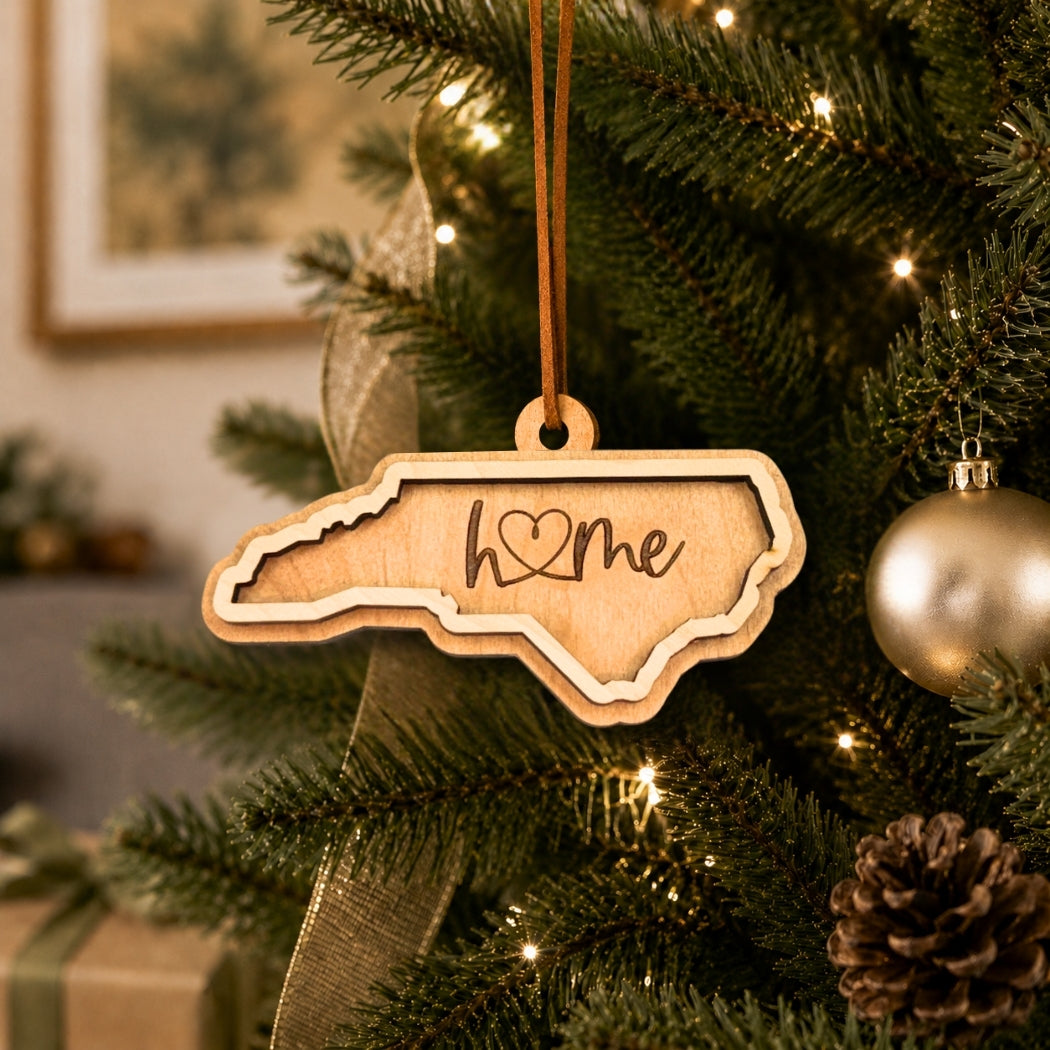 Layered North Carolina “home” ornament hanging on a decorated Christmas tree with warm white lights, pinecones, and gold ornaments.