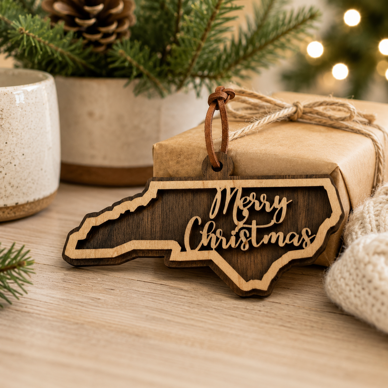 Wooden ornament shaped like North Carolina with 'Merry Christmas' text, tied to a gift box with pine branches and lights in the background.