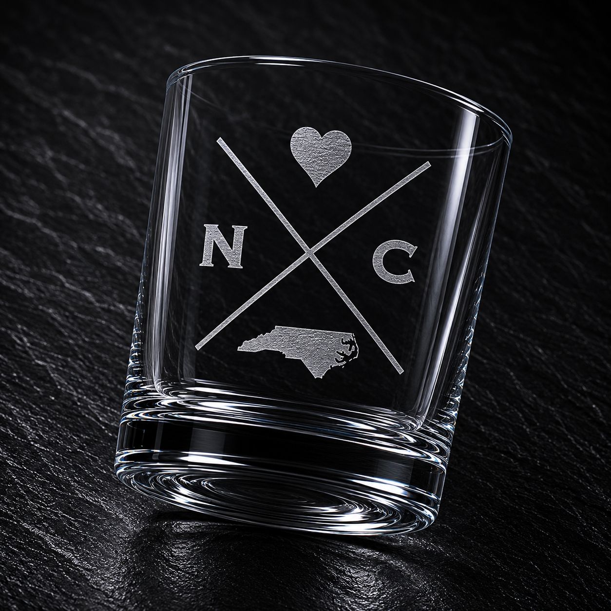 Angled engraved North Carolina whiskey glass featuring frosted North Carolina silhouette and modern crossed-letter design on a dark textured surface.