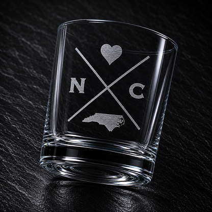 Angled engraved North Carolina whiskey glass featuring frosted North Carolina silhouette and modern crossed-letter design on a dark textured surface.