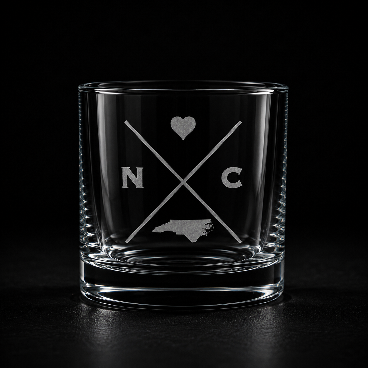 Engraved North Carolina rocks glass with frosted North Carolina silhouette and crossed-letter monogram on a dark background.
