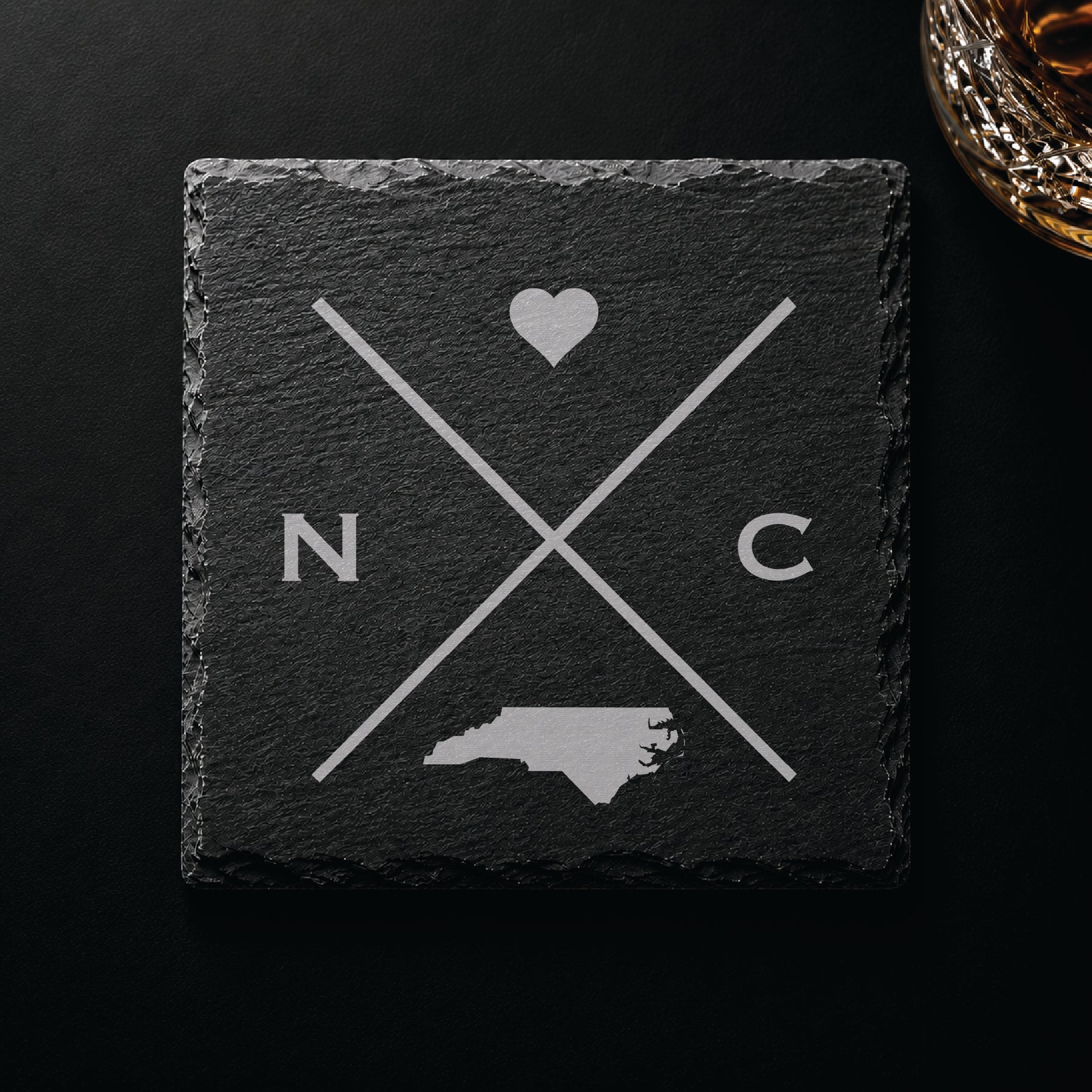 Dark slate coaster with engraved North Carolina crossed-line design, heart icon, and North Carolina silhouette on a dark background.