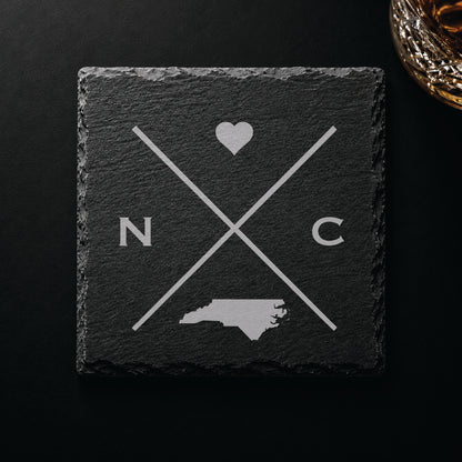 Dark slate coaster with engraved North Carolina crossed-line design, heart icon, and North Carolina silhouette on a dark background.