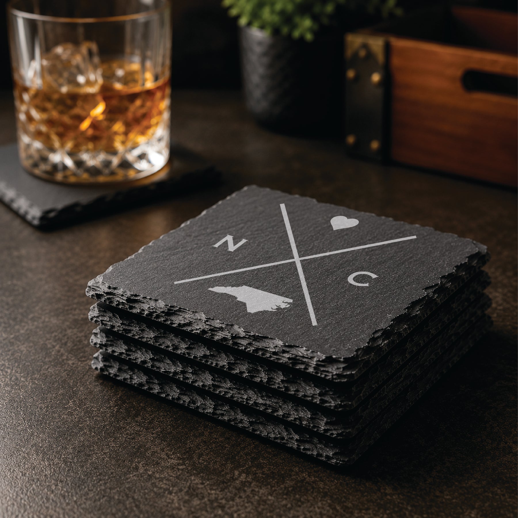 Stack of engraved North Carolina slate coasters in a warm bar-style setting with whiskey glass nearby, featuring a minimalist crossed-line North Carolina design.