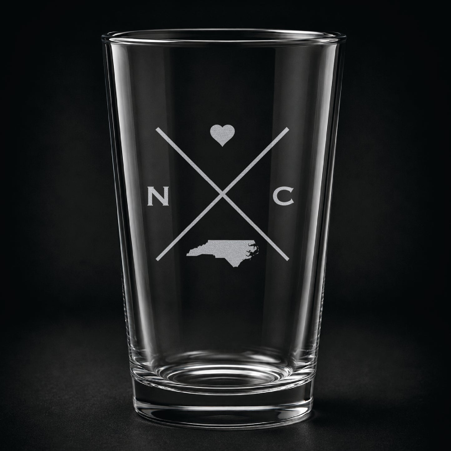 Engraved North Carolina pint glass featuring a minimalist crossed-line design with heart detail and North Carolina silhouette on a dark background.
