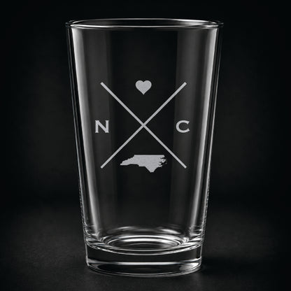 Engraved North Carolina pint glass featuring a minimalist crossed-line design with heart detail and North Carolina silhouette on a dark background.