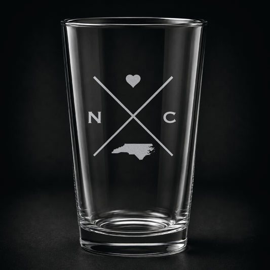 Engraved North Carolina pint glass featuring a minimalist crossed-line design with heart detail and North Carolina silhouette on a dark background.