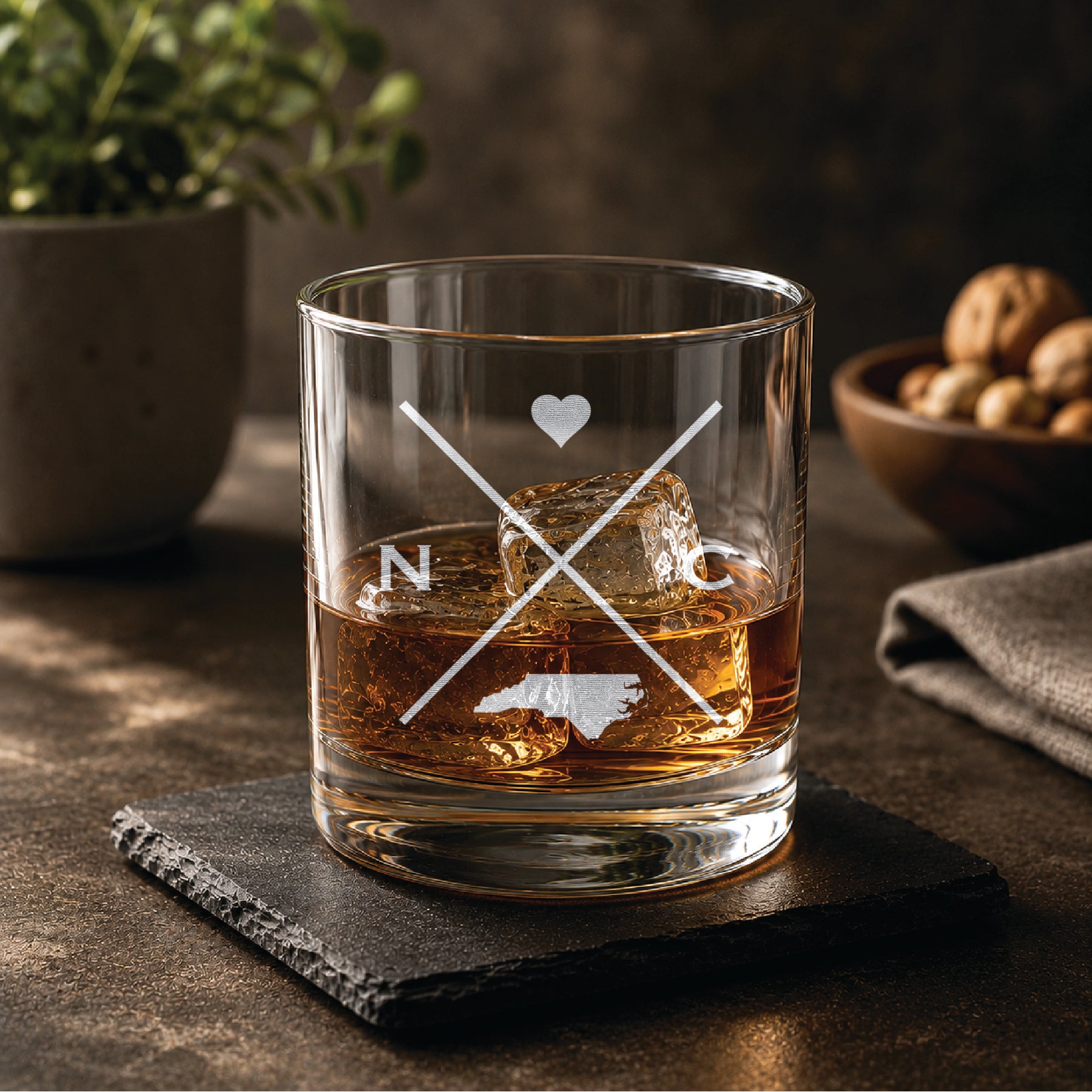 Engraved North Carolina rocks glass with crossed-line monogram design, heart detail, and North Carolina  state silhouette, styled with whiskey and ice on a slate coaster in a warm, upscale bar setting.