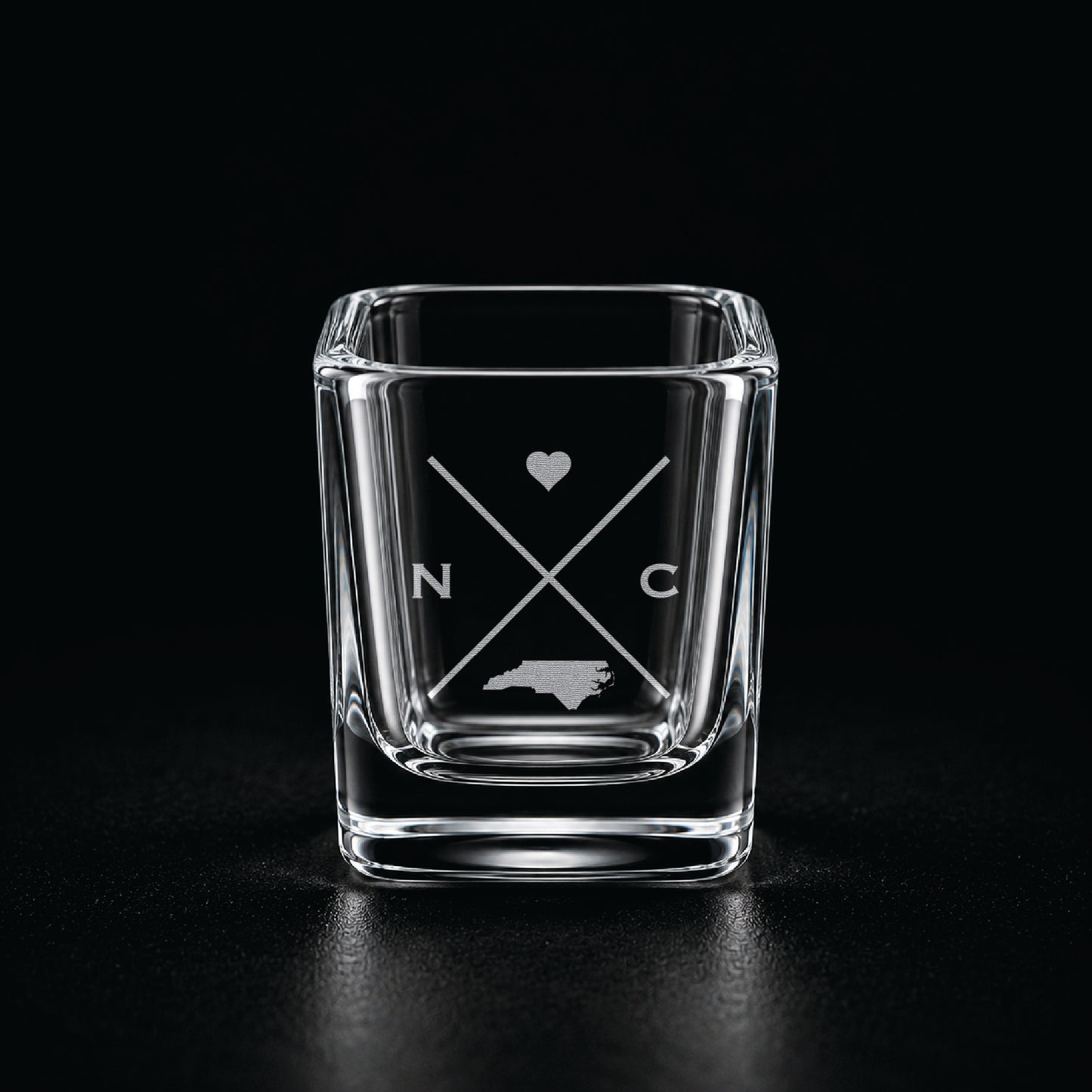 Engraved North Carolina shot glass with modern crossed-line state design and frosted laser engraving on a dark background.