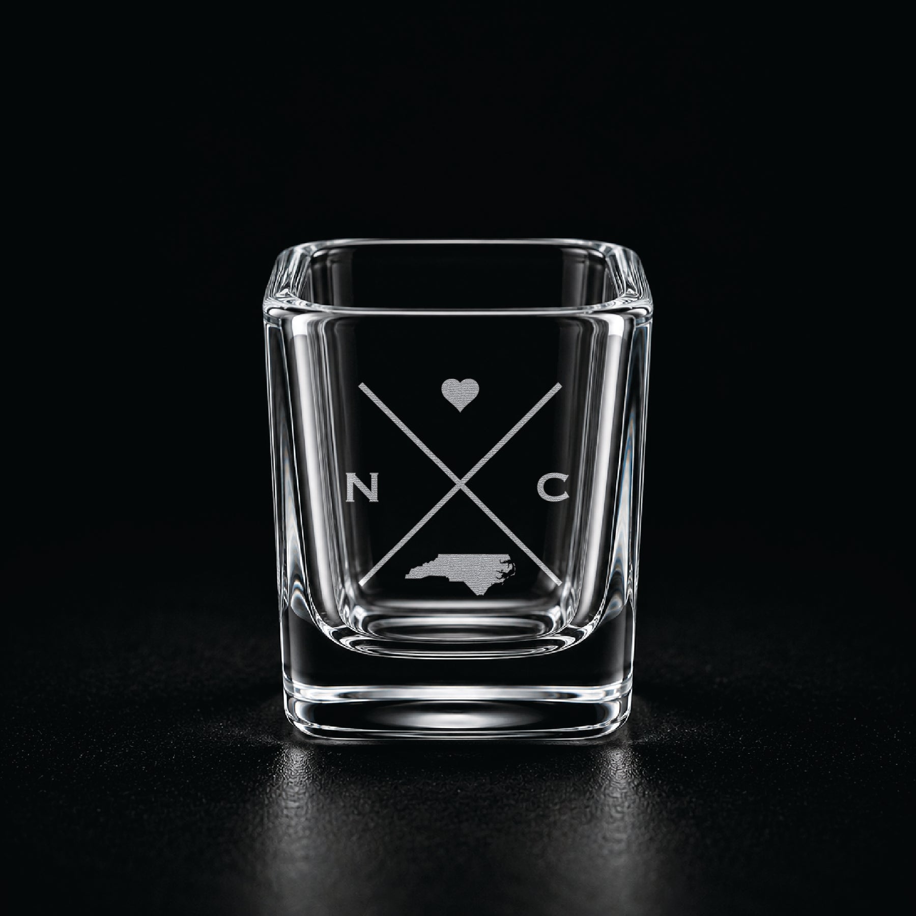 Engraved North Carolina shot glass with modern crossed-line state design and frosted laser engraving on a dark background.