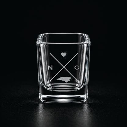 Engraved North Carolina shot glass with modern crossed-line state design and frosted laser engraving on a dark background.