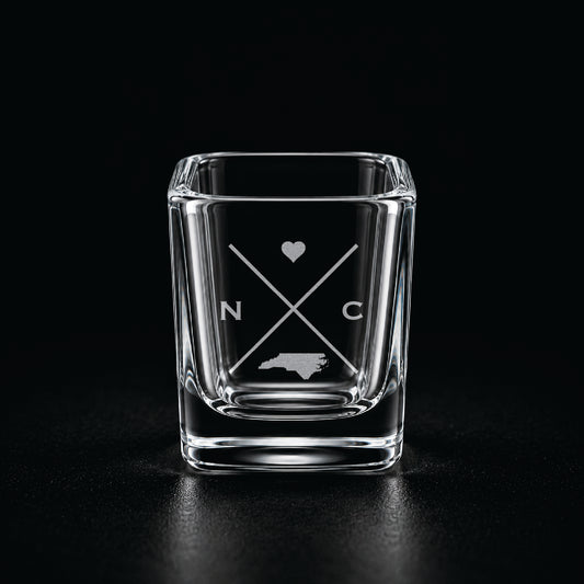 Engraved North Carolina shot glass with modern crossed-line state design and frosted laser engraving on a dark background.