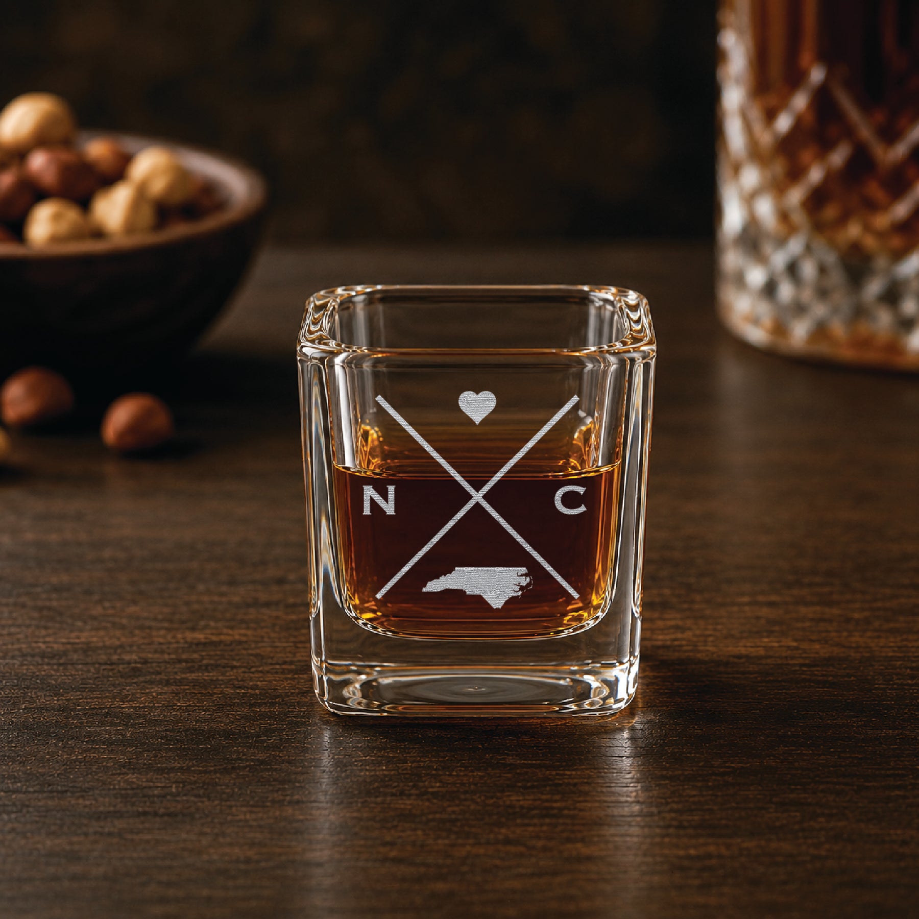 Engraved North Carolina shot glass filled with whiskey in a warm bar-style setting, featuring a minimalist crossed-line design with heart detail and North Carolina silhouette.