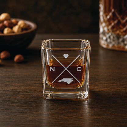 Engraved North Carolina shot glass filled with whiskey in a warm bar-style setting, featuring a minimalist crossed-line design with heart detail and North Carolina silhouette.