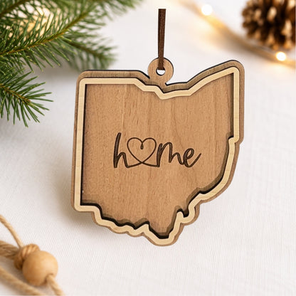 Layered Ohio wood ornament featuring engraved “home” script and faux leather hanger on a soft neutral holiday background with greenery and warm lights.