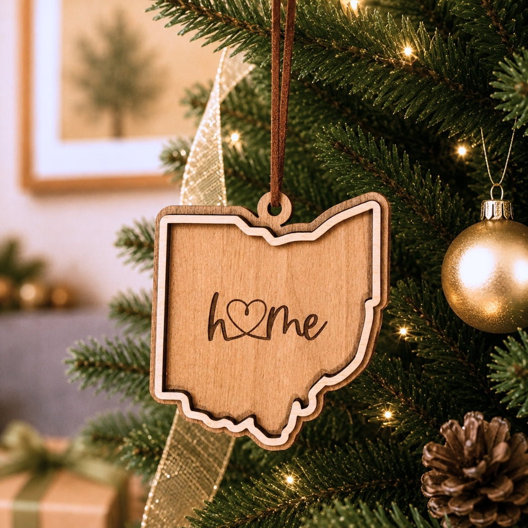Layered Ohio “home” ornament hanging on a decorated Christmas tree with warm white lights, pinecones, and gold ornaments.
