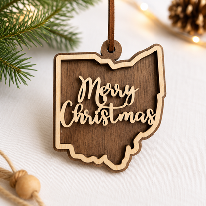 Layered hardwood Ohio Christmas ornament featuring walnut and maple wood with “Merry Christmas” script detail and faux leather hanger styled with evergreen branches and warm holiday lights.