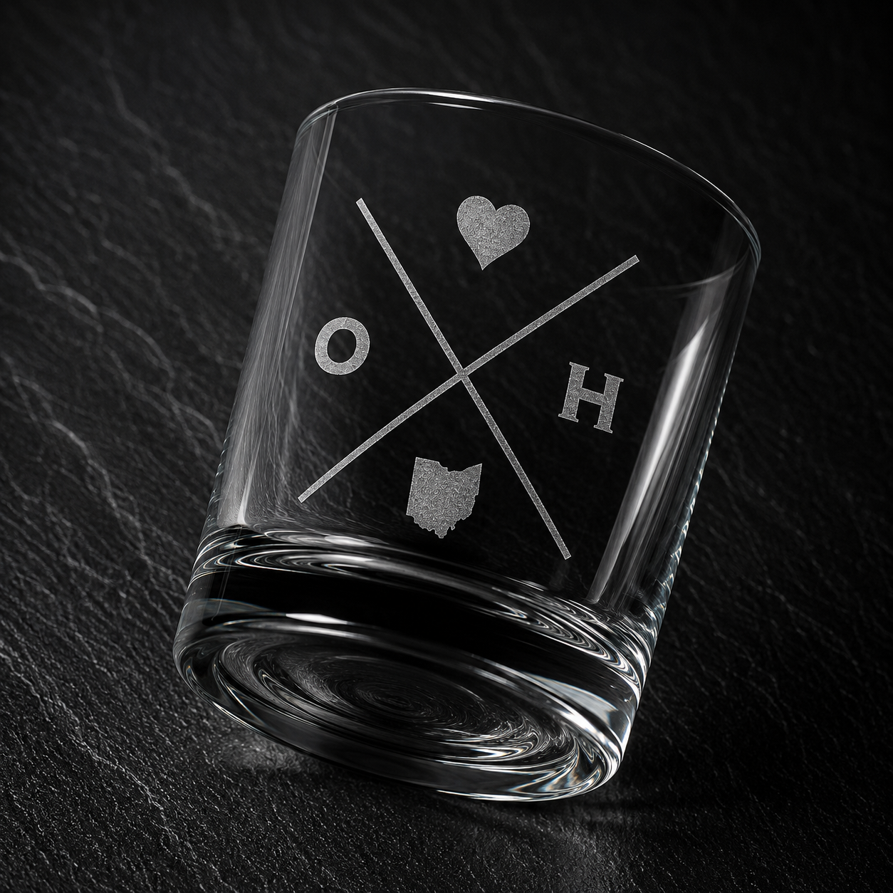 Angled engraved Ohio whiskey glass featuring frosted Ohio silhouette and modern crossed-letter design on a dark textured surface.
