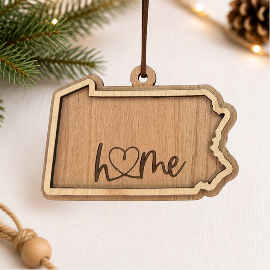 Layered Pennsylvania wood ornament featuring engraved “home” script and faux leather hanger on a soft neutral holiday background with greenery and warm lights.