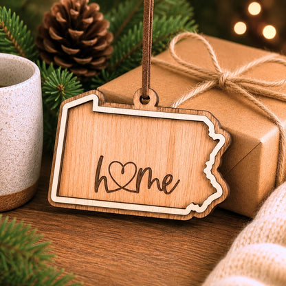 Layered Pennsylvania wood ornament with engraved “home” script displayed beside wrapped holiday gifts, greenery, and warm seasonal décor.