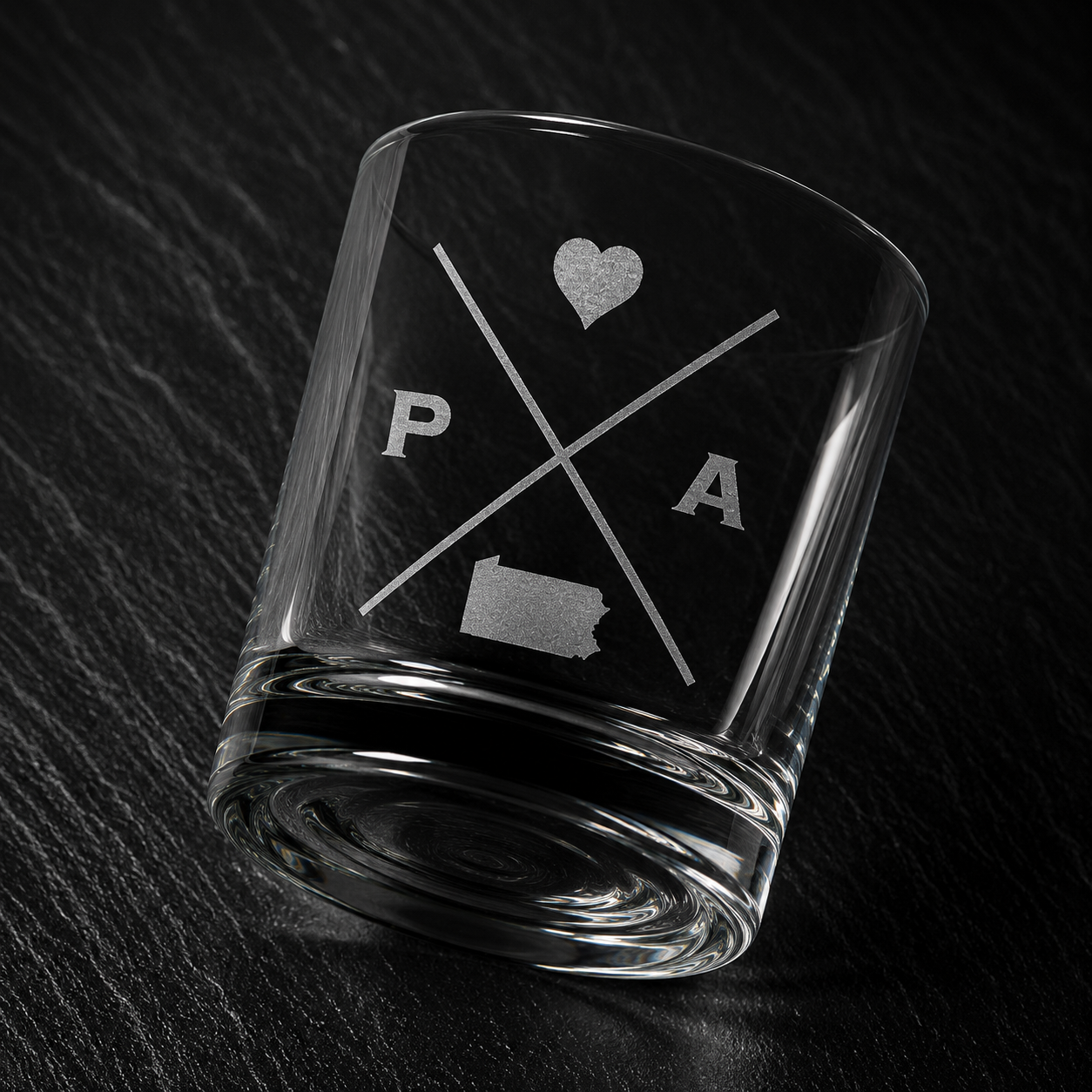 Angled engraved Pennsylvania whiskey glass featuring frosted Pennsylvania silhouette and modern crossed-letter design on a dark textured surface.