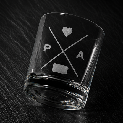 Angled engraved Pennsylvania whiskey glass featuring frosted Pennsylvania silhouette and modern crossed-letter design on a dark textured surface.