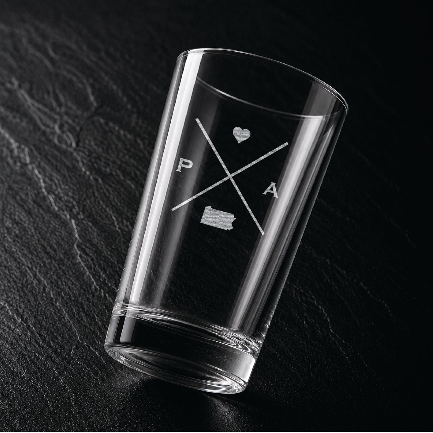 Angled view of engraved Pennsylvania pint glass with modern crossed-line state design and frosted laser engraving on dark textured surface.