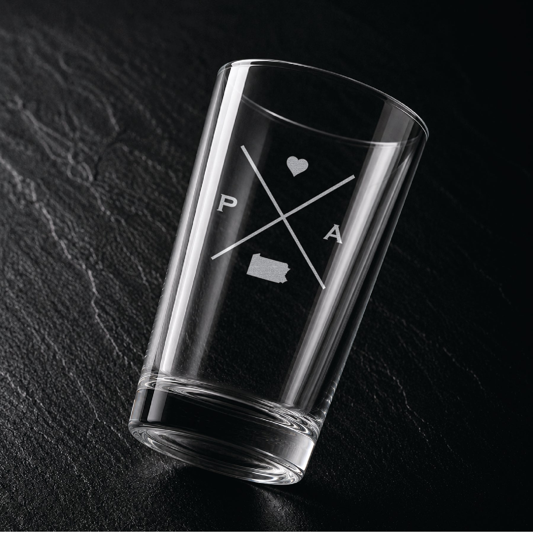 Angled view of engraved Pennsylvania pint glass with modern crossed-line state design and frosted laser engraving on dark textured surface.