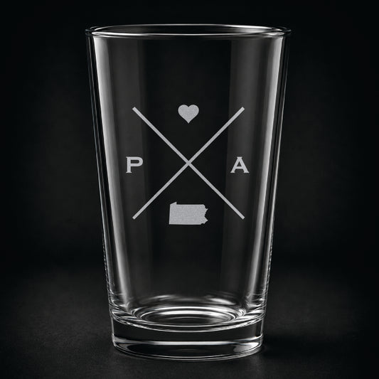 Engraved Pennsylvania pint glass featuring a minimalist crossed-line design with heart detail and Pennsylvania silhouette on a dark background.