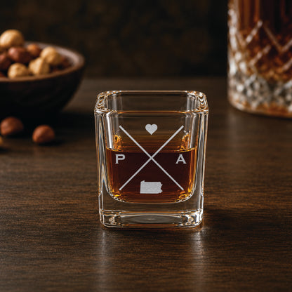 Engraved Pennsylvania shot glass filled with whiskey in a warm bar-style setting, featuring a minimalist crossed-line design with heart detail and Pennsylvania silhouette.