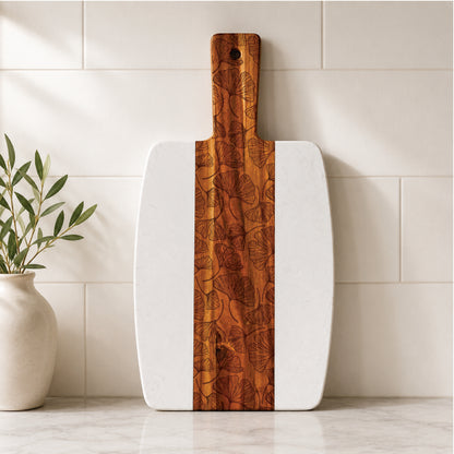 Botanical marble and acacia serving board with engraved ginkgo leaf design displayed upright against a tiled kitchen backsplash beside a ceramic vase with greenery.