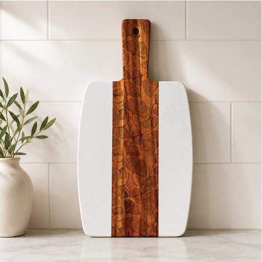 Botanical marble and acacia serving board with engraved ginkgo leaf design displayed upright against a tiled kitchen backsplash beside a ceramic vase with greenery.