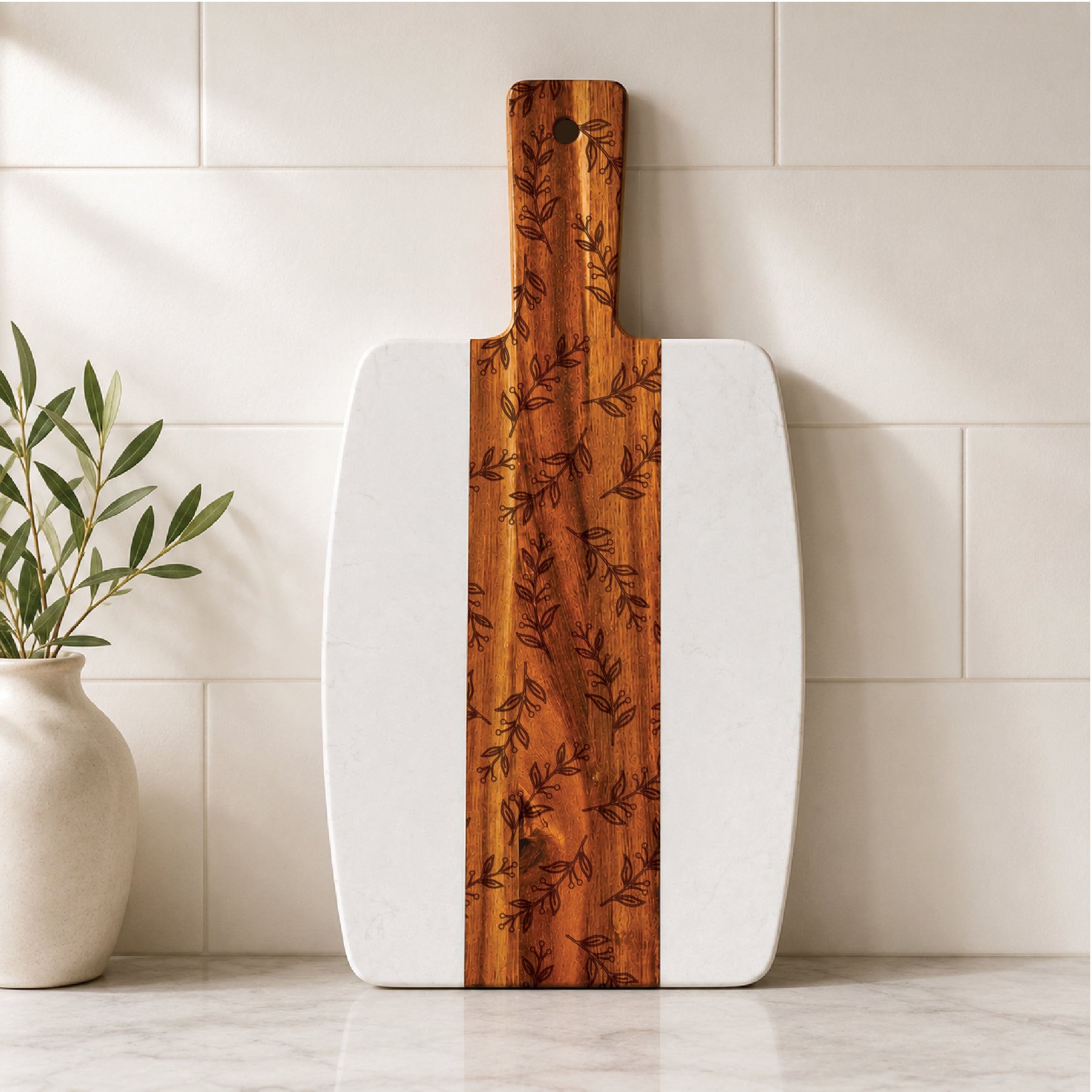 Marble and acacia serving board featuring an engraved botanical pattern, displayed upright on a kitchen countertop against a light tile backsplash.