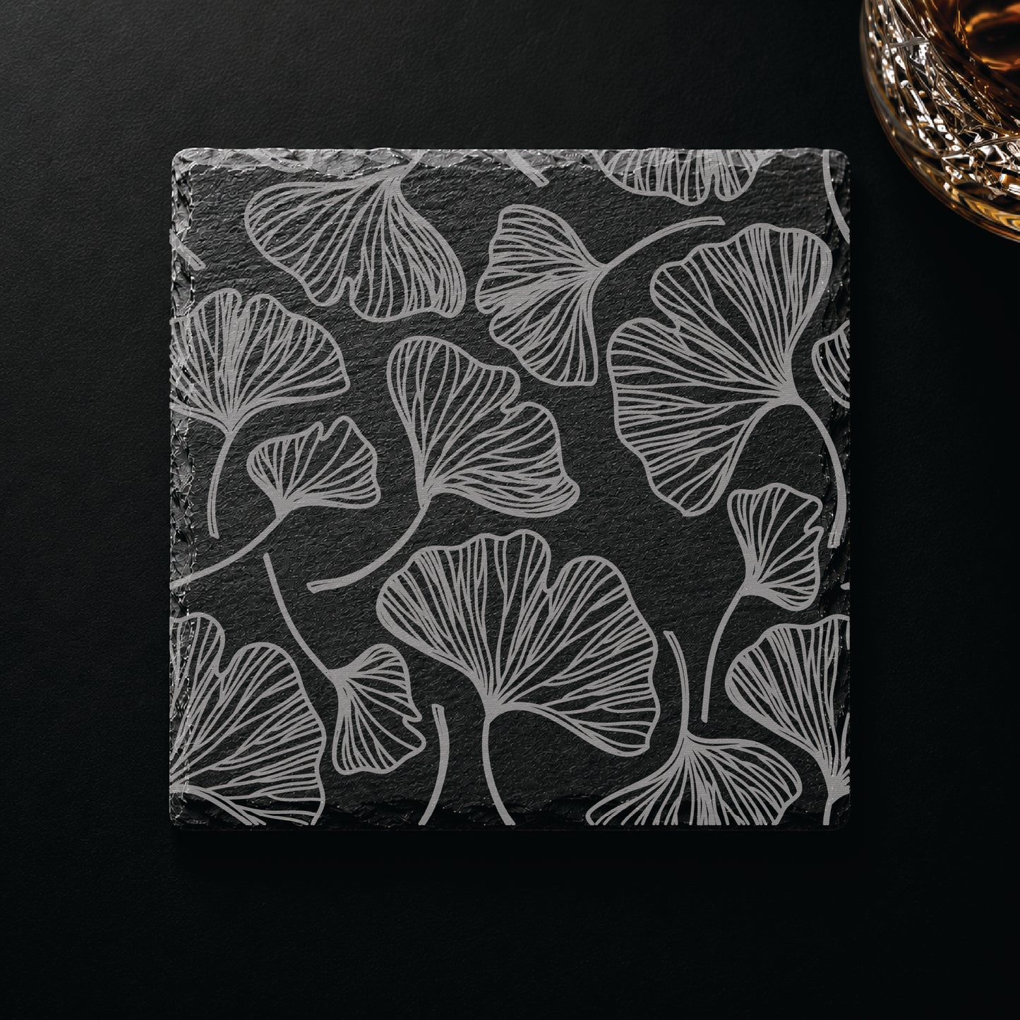 Botanical engraved slate coaster featuring a gingko leaf pattern displayed on a dark tabletop beside a whiskey glass.