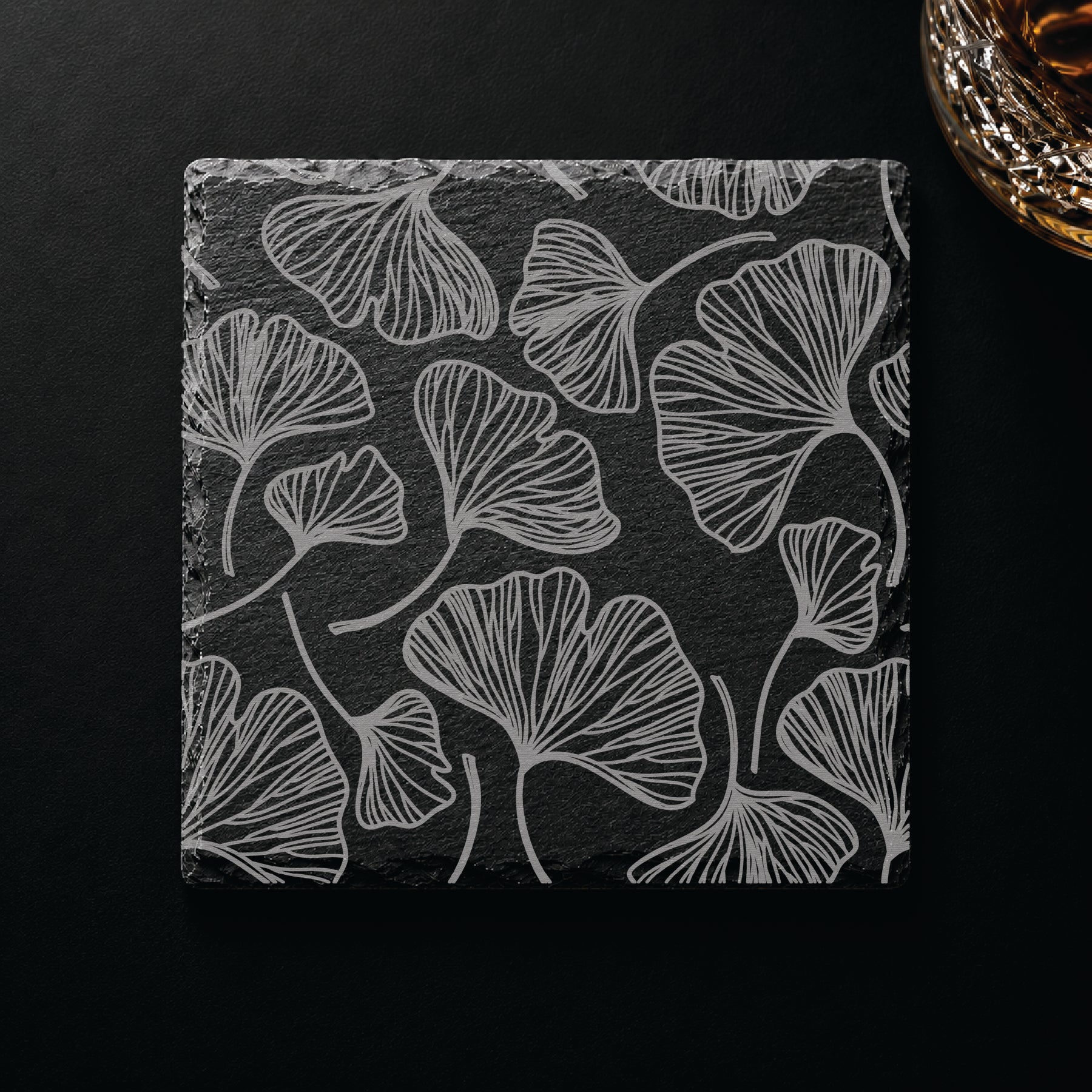 Botanical engraved slate coaster featuring a gingko leaf pattern displayed on a dark tabletop beside a whiskey glass.
