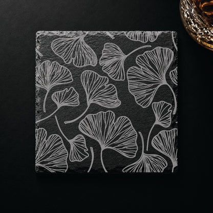 Botanical engraved slate coaster featuring a gingko leaf pattern displayed on a dark tabletop beside a whiskey glass.