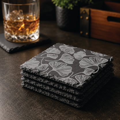 Stacked set of botanical gingko leaf engraved slate coasters on a dark tabletop with whiskey glass and bar décor.
