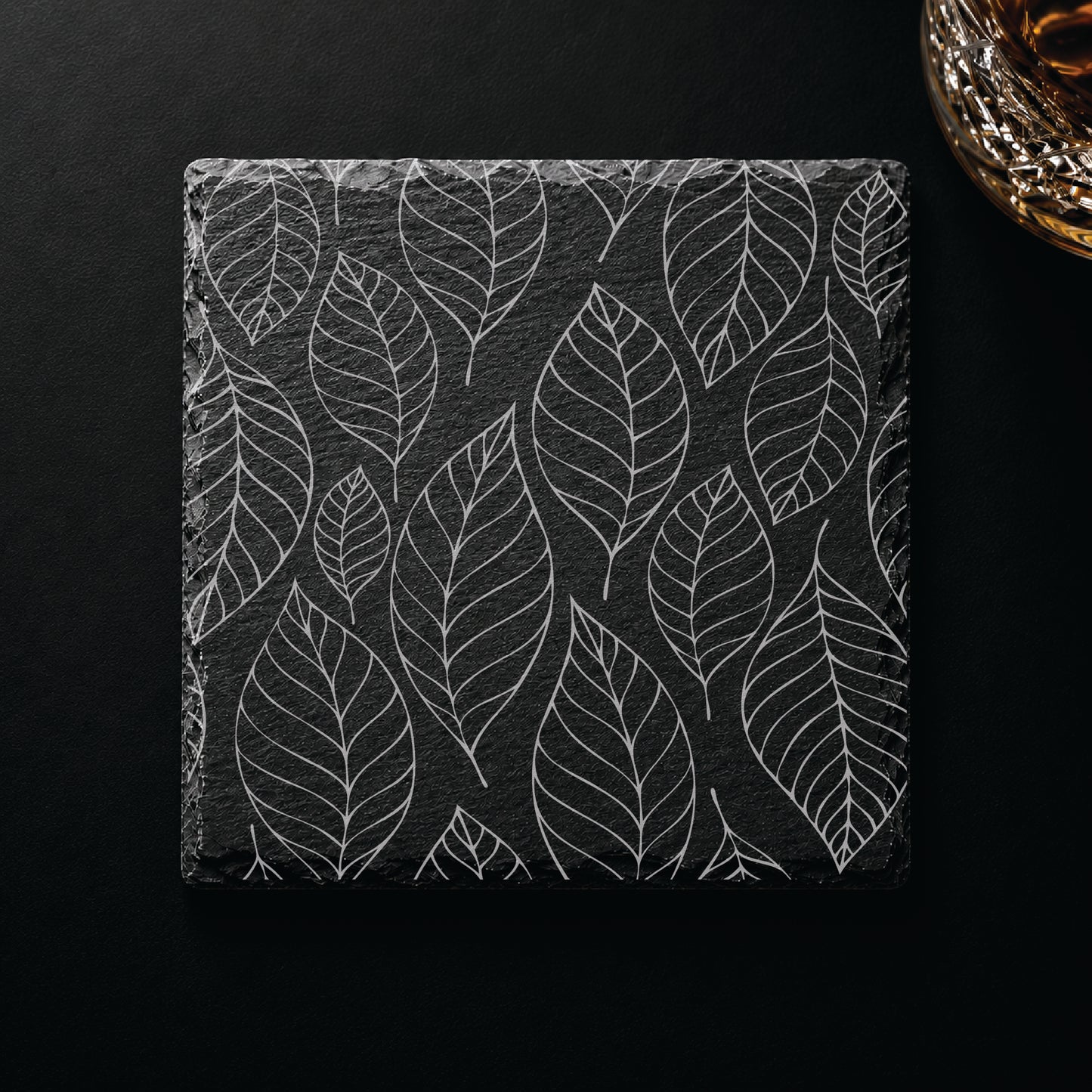 Falling leaves engraved slate coaster displayed on a dark tabletop beside a whiskey glass, featuring a nature-inspired leaf pattern.