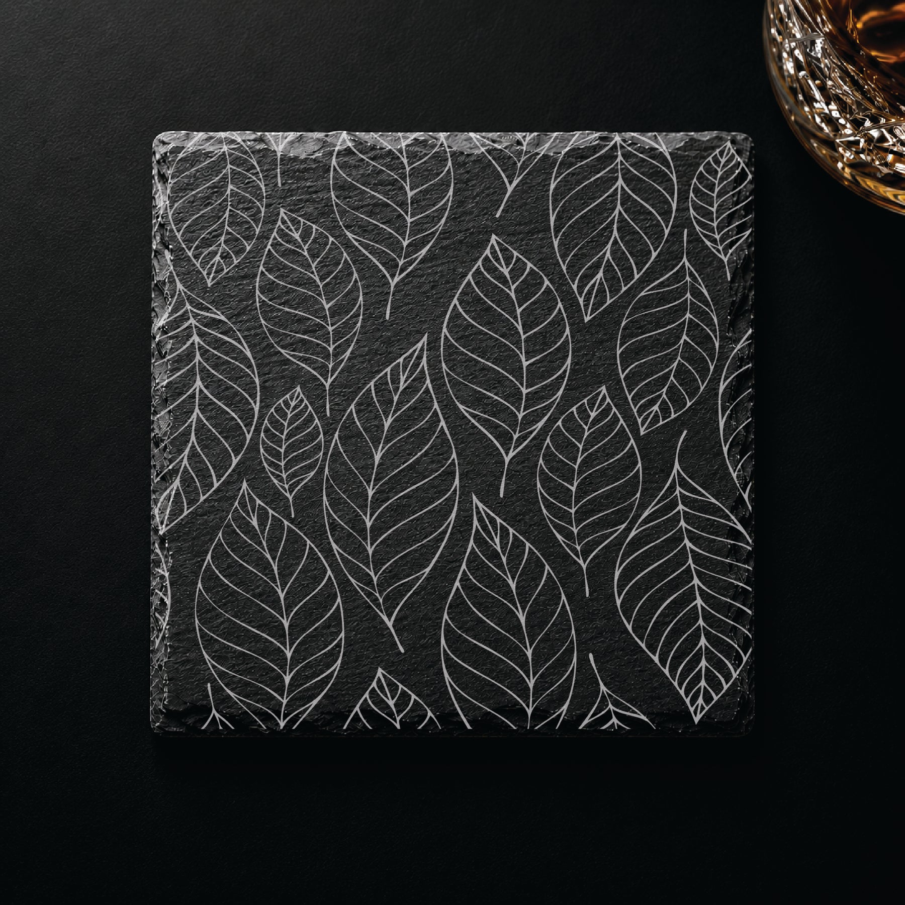 Falling leaves engraved slate coaster displayed on a dark tabletop beside a whiskey glass, featuring a nature-inspired leaf pattern.