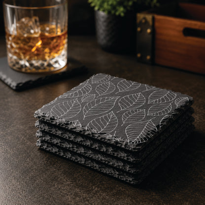 Stacked set of engraved slate coasters featuring a falling leaves design displayed on a dark tabletop with whiskey glass and bar décor.