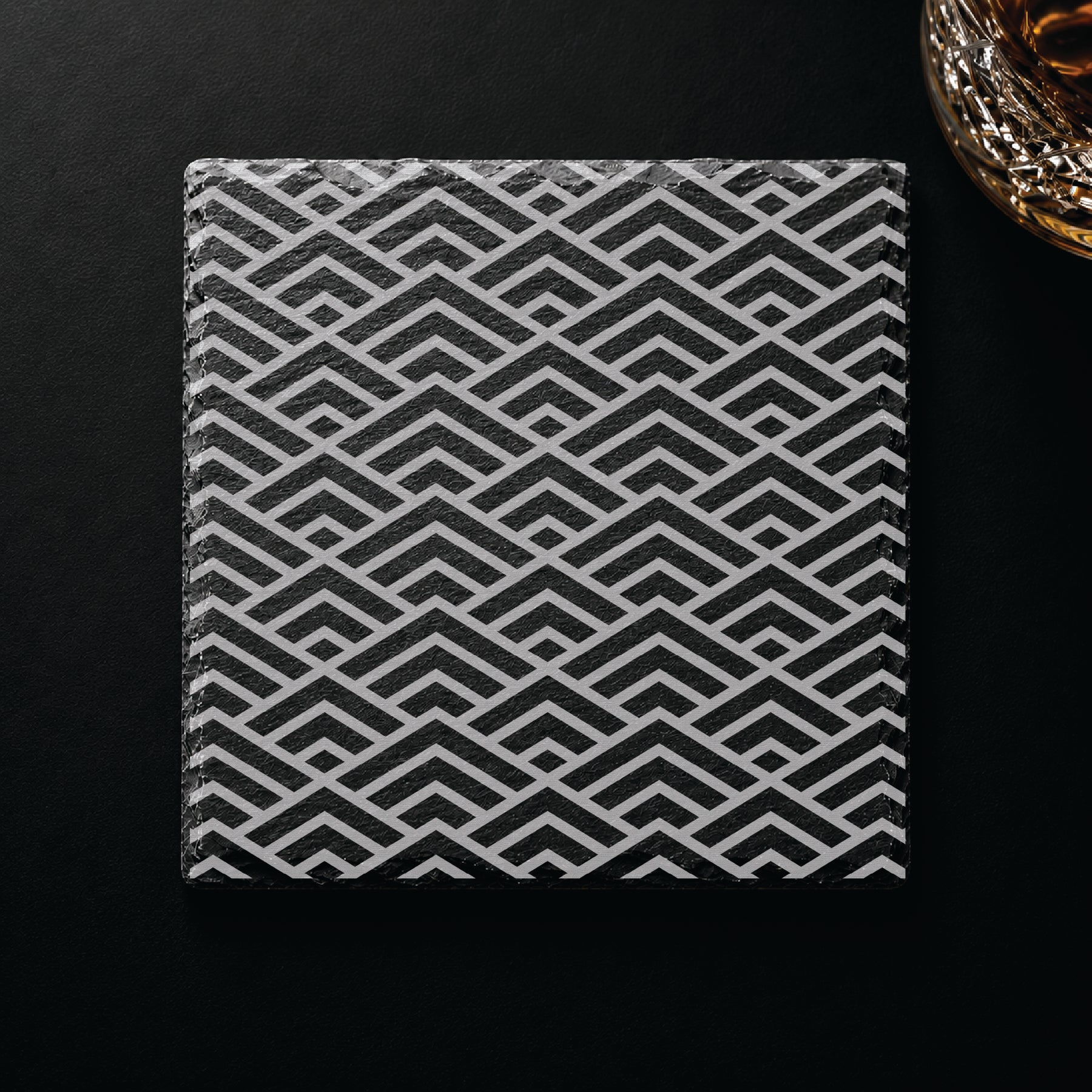 Geometric Peaks engraved slate coaster displayed on a dark tabletop beside a whiskey glass, showcasing a modern geometric design.